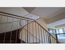 Innenansicht eines Treppenhauses mit gewendelten Treppenläufen und hellen Wänden, eingefasst von einem Metallgeländer. Große Fensterflächen lassen Tageslicht einfallen und geben den Blick nach außen frei.