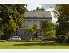 Zweistöckiges Gebäude mit grauer Fassade und Schrägdach, umgeben von Bäumen. Im Vordergrund befindet sich eine Rasenfläche, parkende Autos und ein Zaun.