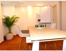 Heller Besprechungsraum mit einem Holztisch, umgeben von weißen Stühlen, einer dekorativen Pflanze in einem Topf links und einem Flipchart im Hintergrund. Der Boden ist mit rotbraunen Fliesen ausgelegt, und die Wände sind weiß gestrichen.