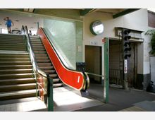 Treppenhaus einer U-Bahn-Station mit einer Treppe links und einer Rolltreppe mit rotem Geländer rechts, flankiert von grünen Fliesenwänden. Im Hintergrund ein Wanduhren, ein Hinweisschild auf barrierefreie Toiletten und ein Verkehrssignal.