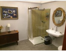 Badezimmer mit einer modernen Dusche in einer Ecke mit goldfarbenen Mosaikfliesen, einem weißen Waschbecken mit darüberhängendem goldenen Spiegel und einer dunklen Holzkommode mit Lampe und Dekoration. Die Wände sind schlicht weiß, der Boden ist mit grauen Fliesen versehen.