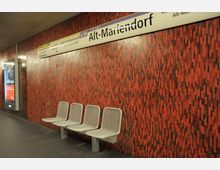 U-Bahn-Station mit roter, gemusterter Wandverkleidung aus kleinen Mosaiksteinen, einer Reihe grauer Metallbänke und einem Stationsschild mit der Aufschrift „Alt-Mariendorf“.