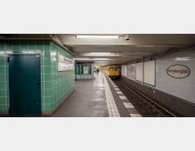 U-Bahnhof mit grün gekachelten Wänden, weißen Namensschildern mit der Aufschrift "Wittenbergplatz" und einem einfahrenden gelben U-Bahn-Wagen auf einem der Gleise. Die Wandflächen gegenüber sind mit weißen Fliesen verkleidet, und eine überdachte Wartezone befindet sich auf der linken Seite des Bahnsteigs.