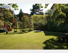 Ein gepflegter Garten mit einer großen Rasenfläche, umgeben von Bäumen und Sträuchern. Im Hintergrund stehen Metallrankhilfen und eine kleine Holztreppe, die zu einer Terrasse führt.