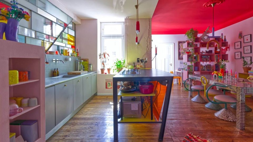 Bright, colorful open-plan kitchen and dining area with a long counter and sink beneath stained-glass style windows, a central island worktable, and wooden plank flooring. The adjoining dining space features a vivid red ceiling, mosaic-tiled table, pastel chairs, open shelving with plants, and framed wall art.