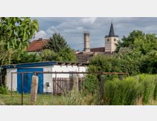 Gartenfläche mit hoher Vegetation und einem kleinen weißen Schuppen mit blauer Tür im Vordergrund; im Hintergrund sind üppige Büsche, Häuser mit Ziegeldächern sowie der Turm einer Kirche mit schwarzem spitzen Dach und Uhr sichtbar.