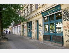 Straßenszene mit einer Hausfassade im Altbaustil, die mit Graffiti bedeckt ist. Links der Gehweg mit Baumschatten und Fußgängern, rechts ein Ladenlokal mit großen blauen Fenstern und einem Schild mit der Aufschrift "Italienische Spezialitäten".
