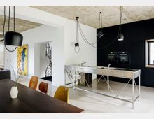 Moderne Küche mit offenem Wohnbereich, geprägt von einer reduzierten Einrichtung. Sichtbetondecke, freistehende Edelstahlküchenzeile, schwarze Wände mit eingebauten Küchengeräten und diskreter Beleuchtung; im Hintergrund eine Wendeltreppe und ein Kunstgemälde.