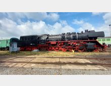 Eine schwarze Dampflokomotive mit roten Rädern und Gestellen steht auf einem Bahngleis im Freien. Im Hintergrund sind weitere, grün lackierte Eisenbahnwagen zu sehen, sowie ein kleines Gebäudeelement und ein wolkiger Himmel.