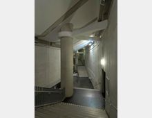 Treppenbereich in einem modernen, unterirdischen Gebäude mit Betonwänden, metallischen Geländern und einer zentralen Betonsäule. Im Hintergrund führen Treppenstufen in eine höhere Ebene, und an den Wänden sind Leuchten angebracht.