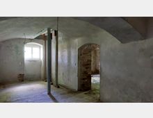 Ein leerer Raum mit gewölbter Decke und sichtbaren Putzschäden an den Wänden. Im Hintergrund befindet sich ein kleines Fenster mit Holzrahmen und ein gemauerter Türbogen, der in einen weiteren Raum mit freiliegendem Ziegelwerk führt.