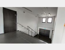 Ein schlichtes Treppenhaus mit Metallgeländern, weißen Wänden und grauem Bodenbelag. Oben befindet sich eine geschlossene Tür, unten zwei große Doppeltüren mit einem grün beleuchteten Notausgangsschild darüber; an der Wand sind zwei kleine Fenster.