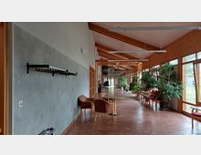 Ein heller, länglicher Flur mit großen Fenstern auf der rechten Seite, die Tageslicht hereinlassen. Der Raum ist mit beigen Holz- und Metallträgern gestaltet, verfügt über geflieste Böden, mehrere Sitzgelegenheiten, Topfpflanzen und eine Garderobenstange an der linken Wand.