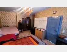 Ein einfach eingerichtetes Zimmer mit gemusterten Tapeten, einem Doppelbett, einem Kleiderschrank, einem Holzofen und einer kleinen Küchenecke mit Waschbecken. Ein auffälliger Kronleuchter hängt von der Decke, der Boden ist mit Fliesen und einem Teppich bedeckt, und eine blaue Tür führt aus dem Raum.