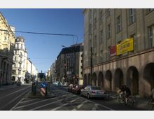 Straßenszene in einer Stadt mit klassischen und modernen Gebäuden, Straßenbahngleisen, Autos und Radfahrern. Auf der rechten Seite befinden sich Gebäude mit Arkaden und einem Werbeschild für Bürovermietung.