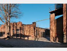 Brick industrial courtyard with cobblestone paving, surrounded by low red-brick buildings with metal doors and arched windows. A tall brick tower with a small platform and railings stands on the right, with a leafless tree casting shadows across the walls under a clear blue sky.