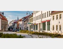 Straßenszene in einer Kleinstadt mit mehrstöckigen Wohn- und Geschäftshäusern, teils mit Schaufenstern und roten Dächern. Im Vordergrund ein Kreisverkehr mit Blumenbeeten, dahinter Autos, Fußwege und Straßenschilder; im Hintergrund ein Kirchturm.