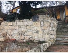 Steinmauer mit verwitterten Steinstufen, die zu einem gelben Haus mit Flachdach und großer Fensterfront im Hintergrund führen. Im Vordergrund sind Pflanzen, Kies und ein eingelassenes Relief einer Gesichtsskulptur sichtbar.