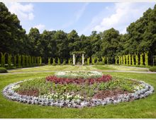 Ein symmetrischer Garten mit kreisförmigem Blumenbeet im Vordergrund, gesäumt von bunten Pflanzen und umgeben von gepflegtem Rasen. Im Hintergrund sind eine Pergola mit Säulen sowie grüne Rankbögen und eine Baumreihe zu sehen.