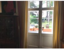 Ein Innenraum mit einem weißen Doppelfenster, das auf eine Terrasse und einen Garten mit Bäumen und Pflanzen hinausblickt. Die Fenster sind von Vorhängen in Gelb- und Rottönen eingerahmt, und ein dunkler Holzschrank steht links daneben.