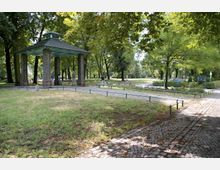 Parklandschaft mit einem kleinen Pavillon aus Backsteinsäulen und einem grünen Dach, umgeben von gepflasterten Wegen, Rasenflächen und Bäumen. Im Hintergrund sind weitere Parkelemente wie Blumenbeete und Sitzbänke zu sehen.