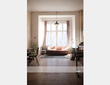 Wohnzimmer mit hellen Wänden und Holzboden, im Mittelpunkt ein großes Fenster mit rosa Vorhängen. Vor dem Fenster steht ein rosa Sofa auf einem Teppich, flankiert von Pflanzen und einem Beistelltisch, mit einer hängenden Deckenleuchte darüber.