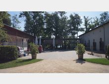 Ein gepflasterter Hof mit einem hölzernen Pavillon und geschlossenen Sonnenschirmen, umgeben von zwei Gebäuden; links ein Holzgebäude, rechts ein weißes Gebäude mit Stalltoren. Im Hintergrund erstreckt sich eine Baumreihe, durch die Wasser sichtbar ist.