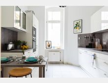 Helle, moderne Küche mit weißen Schränken, grauer Arbeitsplatte und einem zentralen Fenster, das viel natürliches Licht hereinlässt. Rechts befindet sich eine Wand mit Küchenutensilien, links ein kleiner Esstisch mit gedeckten Plätzen, und im Raum sind farbige Dekoelemente wie Zitronen und Pflanzen platziert.