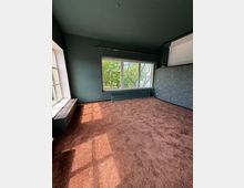 Leerer Innenraum mit dunkelgrünen Wänden, braunem Teppichboden und großen Fensterflächen, die auf Bäume im Außenbereich blicken. Links befindet sich eine Fensterbank mit eingelassenen Sitz-/Heizkörperverkleidungen, rechts eine Wand mit blau gemusterter Tapete.