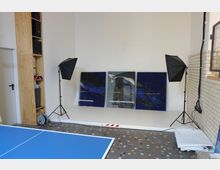 Ein Raum mit einem kleinen, erhöhten Podest, auf dem drei blaue Gemälde nebeneinander aufgestellt sind, flankiert von zwei Studiolampen mit schwarzen Softboxen. Im Vordergrund ein Tischtennistisch, rechts helle Fliesen und links ein Holzregal mit Geräten und Gegenständen.