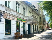 Straßenszene mit einer historischen, verzierten weißen Hausfassade, die mehrere Geschäfte beherbergt, darunter ein Sanitätshaus namens "Schadock". Rechts verläuft eine gepflasterte Straße mit Fußgängern und Bäumen, die Schatten spenden.