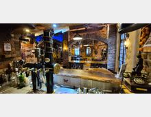 Innenansicht einer rustikalen Bar bzw. Kneipe mit freiliegenden Backsteinwänden, Rundbögen und warmem Deckenlicht. Im Vordergrund steht ein Holztresen mit Zapfanlage und Flaschen, dahinter ein Sitzbereich mit Holztischen und -stühlen sowie dekorativen Vintage-Objekten.