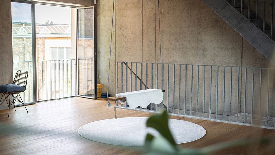 Loftartiger Innenraum mit Sichtbetonwänden, Holzdecke und Holzfußboden, in dem eine hängende Wiege über einem runden Teppich befestigt ist. Links führt eine große Glas-Schiebetür auf einen Balkon, rechts verläuft eine Metalltreppe entlang der Wand.