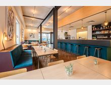 Modernes Café mit Holztischen, blauen Polsterbänken und einer langen Theke mit Barhockern. Die Einrichtung umfasst helle Wände mit abstrakter Kunst, hölzerne Bodenfliesen und dezente Beleuchtung durch hängende Lampen.