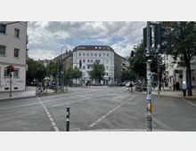 Straßenkreuzung in einer Stadt mit mehrstöckigen Wohngebäuden, Fuß- und Fahrradwegen sowie mehreren Verkehrsampeln. Im Hintergrund stehen Bäume und parkende Fahrräder, während Autos und eine Radfahrerin die Straße überqueren.