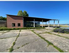 Ein modernes Backsteingebäude mit großen Glasfronten und Flachdach steht neben einer überdachten offenen Fläche mit Säulen. Im Vordergrund ist ein gepflasterter Hof mit vereinzeltem Grasbewuchs, im Hintergrund Schilf und ein See unter klarem Himmel sichtbar.