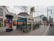 Städtisches Straßenbild am Kurfürstendamm in Berlin mit einem Eingang zur U-Bahn-Station "U Kurfürstendamm", umgeben von Geschäftsfassaden, einem Werbesäulenplakat und Fahrrädern. Im Hintergrund sind ein Einkaufszentrum, ein Hotel und die Turmruine der Kaiser-Wilhelm-Gedächtniskirche sichtbar, ebenso wie Baukräne und Straßenlaternen.