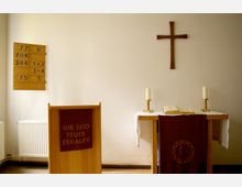 Ein schlichter Kirchenraum mit einem Holzkreuz an der Wand, einem Altar mit weißer Tischdecke, zwei Kerzenleuchtern und einer geöffneten Bibel. Im Vordergrund steht ein Redepult mit dem Schriftzug „Ihr seid teuer erkauft“, daneben eine Tafel mit Liednummern.