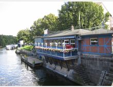 Backstein-Gebäude mit blauen Fachwerkelementen und einer Terrasse mit Blumenbeeten, direkt am Ufer eines Flusses. Im Hintergrund sind ein Anlegesteg und ein weißes Passagierschiff zu sehen, umgeben von Bäumen und Grünflächen.