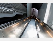 Eine abwärtsführende Rolltreppe in einem modernen, minimalistischen Innenraum mit glatten Metalloberflächen und Lichtstreifen entlang der Seiten. Im Hintergrund sind Treppen und eine Wand mit Metallgeländern sichtbar.