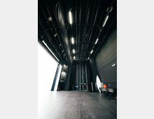 Blick auf die Bühne eines modernen Theater- oder Veranstaltungssaals mit schwarzem Boden, hohen schwarzen Wänden und zahlreichen Bühnenvorhängen. Deckenbeleuchtung sowie Bühnentechnik wie Kabel, Scheinwerfer und Transporteinrichtungen sind sichtbar.