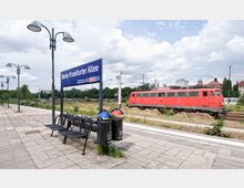Bahnhof Berlin Frankfurter Allee mit einem blauen Stationsschild und Sitzbänken im Vordergrund, im Hintergrund ein roter Zug der Deutschen Bahn sowie Gleise, umgeben von Bäumen und städtischen Gebäuden. Straßenlampen und Abfalleimer sind entlang des gepflasterten Bahnsteigs zu sehen.