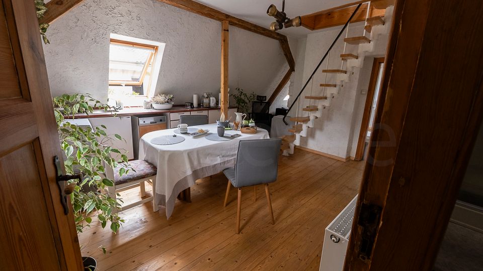 Heller Dachgeschossraum mit Holzboden, sichtbaren Deckenbalken und einem Esstisch mit weißer Tischdecke in der Mitte. Links eine kleine Küchenzeile unter einem Dachfenster, rechts eine schmale Holztreppe mit Geländer zu einer oberen Ebene.
