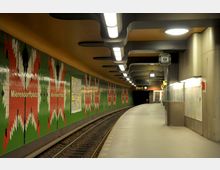 U-Bahnhof mit modernem Design; grüne Wandfliesen mit roten und weißen Musterelementen, auf denen "Mierendorffplatz" steht, leuchtende Deckenlampen und geschwungene Bahnsteigabdeckung. Im Vordergrund Gleise, im Hintergrund ein Fahrplan und eine Rolltreppe.