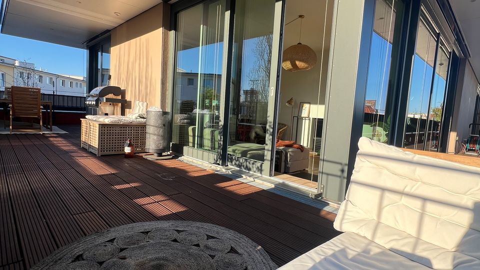 Große Dachterrasse eines modernen Wohngebäudes mit Holzdeck, breitem Dachüberstand und bodentiefen Glasschiebetüren entlang der Fassade. Auf der Terrasse stehen Sitzmöbel und ein runder Outdoor-Teppich; im Hintergrund sind umliegende Gebäude unter blauem Himmel zu sehen.