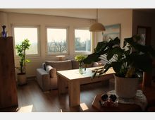 Wohnzimmer mit hellen Holzböden, einer grauen Couch und einem Holztisch, der mit einer Vase mit Blumen dekoriert ist. Große Fenster lassen Tageslicht herein, und im Raum sind grüne Zimmerpflanzen sowie eine Hängelampe sichtbar.