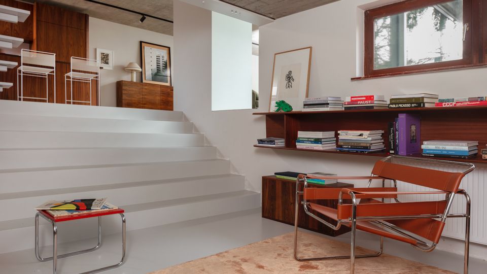 Modern split-level living room with wide white steps leading to a raised seating area, exposed concrete ceiling, and built-in wood shelving filled with books beneath a small window. Minimalist furniture includes chrome-framed chairs and a low table on a large beige rug.