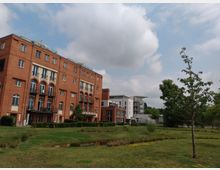 Wohngebäude aus rotem Backstein mit großen Bogenfenstern und Balkonen, flankiert von einem moderneren weißen Gebäude im Hintergrund. Im Vordergrund befindet sich eine Wiese mit kleinen Bäumen und einem schmalen Wasserlauf, umgeben von einer parkähnlichen Landschaft unter einem bewölkten Himmel.