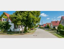 Gepflasterte Wohnstraße in einer ländlichen Gegend mit Einfamilienhäusern, roten Ziegeldächern und gepflegten Vorgärten, flankiert von Bäumen und Hecken unter klarem, blauem Himmel. Ein großes, grün bewachsenes Haus steht im Vordergrund.