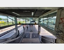 Blick von einer überdachten Treppenanlage auf einen modernen Bahnhof mit mehreren offenen Bahnsteigen. Im Vordergrund sind Glaswände, Fahrbahnmarkierungen und ein Informationsschild zu sehen, während die Bahnsteige mit wartenden Fahrgästen von Zügen umgeben sind.